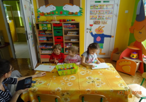 Troje dzieci i jedna osoba dorosła siedzą przy stoliku. Dzieci układają mozaikę.